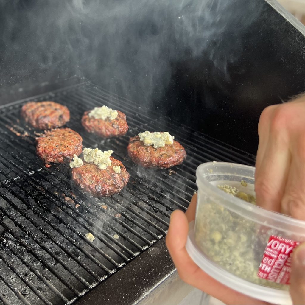Hawks wings Mushroom burgers grilling while someone is adding blue cheese.