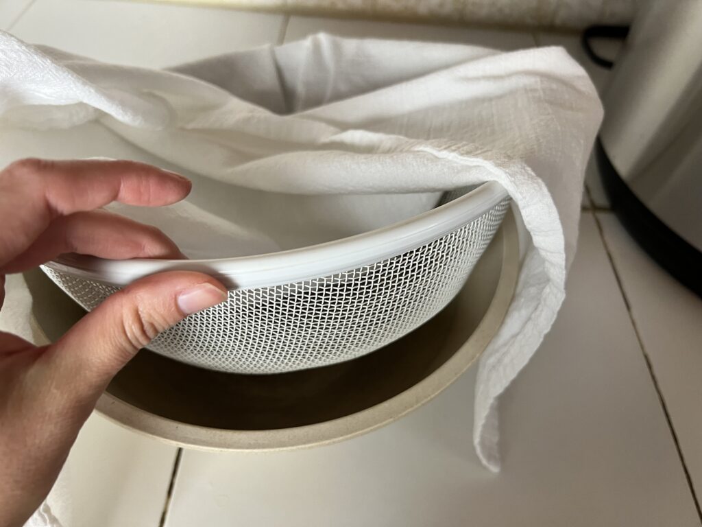 A flour sack towel over a mesh strainer and mixing bowl.