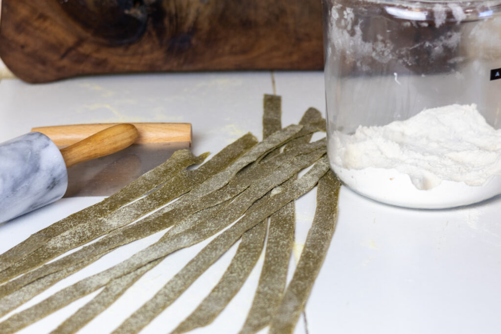 Fresh nettle pappardelle pasta next to flour, bench scraper, and rolling pin.