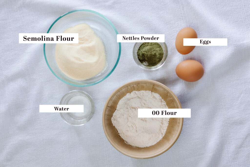 A flat lay of ingredients to make nettles pasta: semolina flour, 00 flour, nettles powder, eggs, and water.