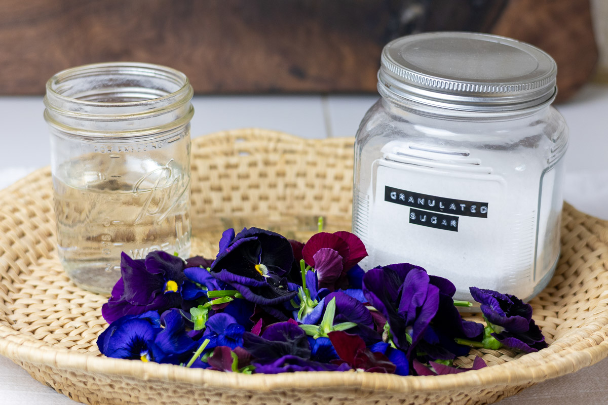 A pile of dark pansies in front of a glass of water and sugar on a basket.