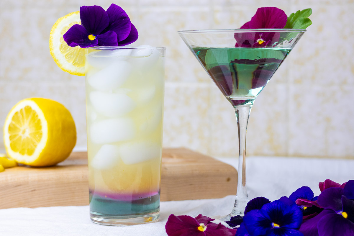 Two drinks, a lemonade and a cocktail with pansy syrup and flowers.