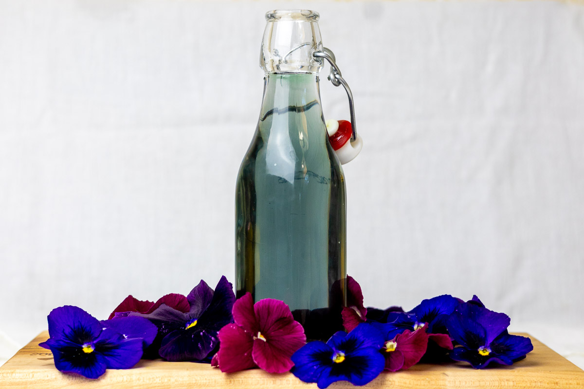 a flip top jar of dark purple syrup with dark pansy flowers on a wooden board.