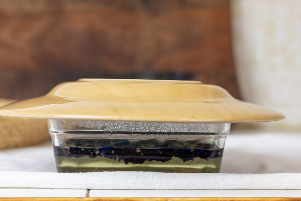 A beige plate on top of a glass container with petals steeping.