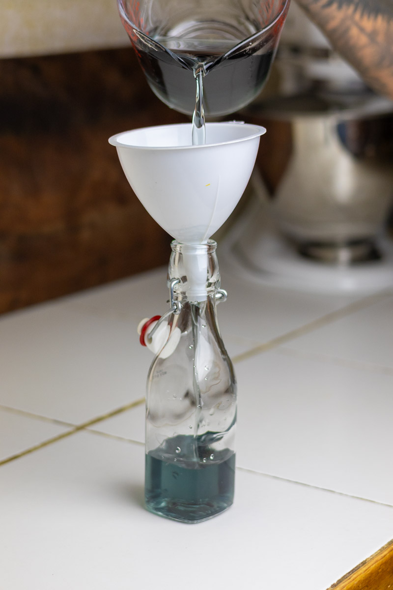 pouring pansy syrup in a swing top bottle through a funnel on white table.