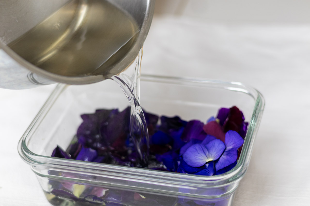 A silver pot pouring syrup over petals in glass container. 