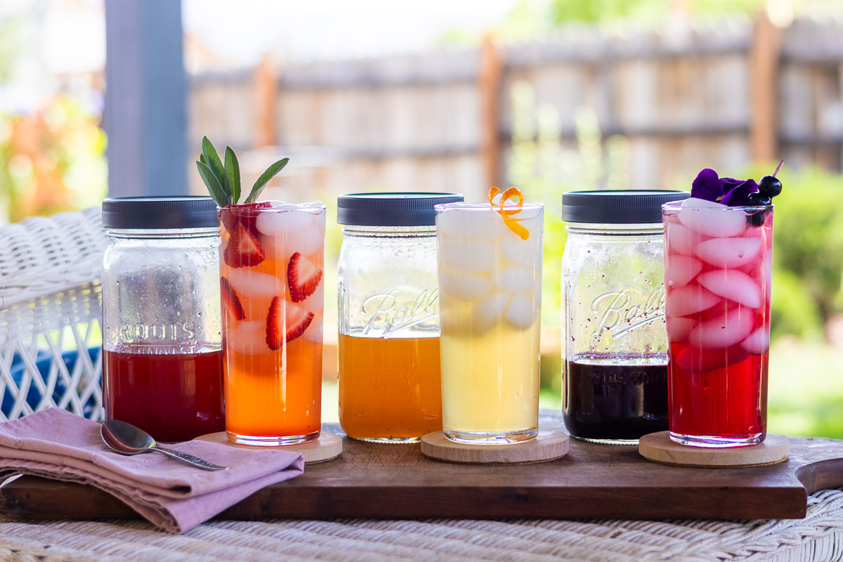 Strawberry, orange and blueberry shrub syrups with three garnished drinks ready to serve.