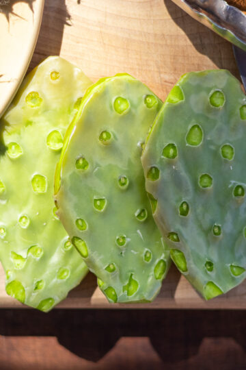 three despined nopales on wood cutting board next to paring knife and tongs.