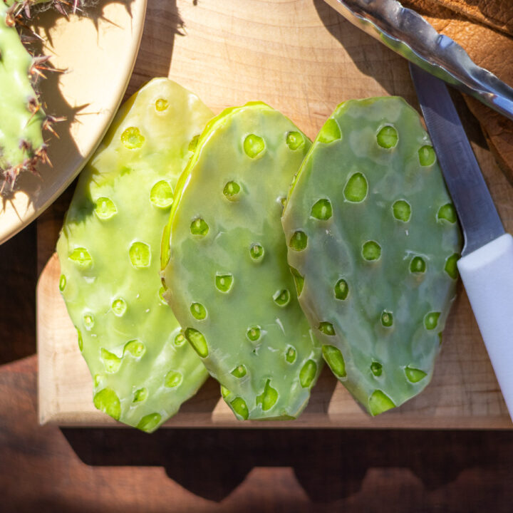 three despined nopales on wood cutting board next to paring knife and tongs.