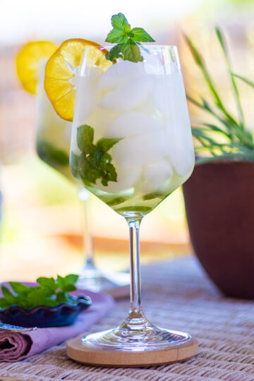 A Hugo Spritz cocktail, napkin, spoon, and fresh mint on a white wicker table.