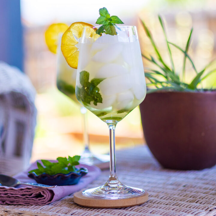 A Hugo Spritz cocktail, napkin, spoon, and fresh mint on a white wicker table.