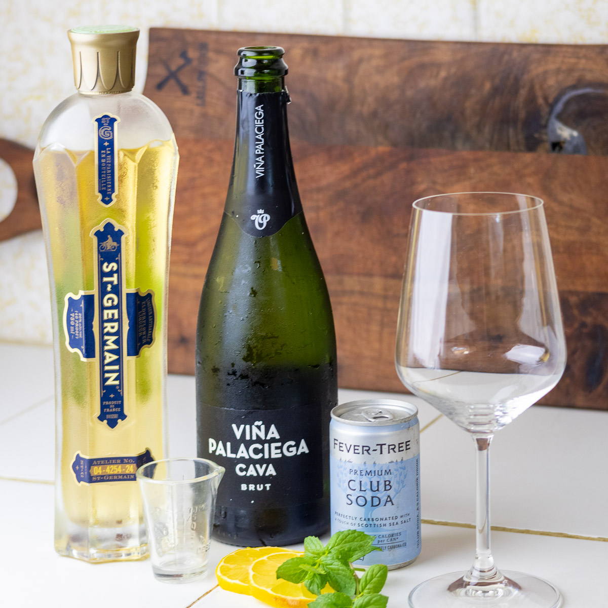 Ingredients for a Hugo Spritz cocktail including St. Germain, Cava, club soda, lemon, mint, shot glass, and a wine glass.