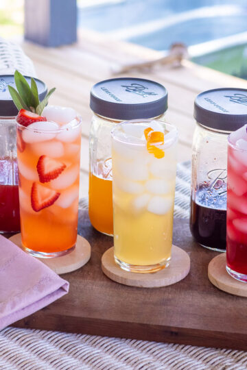 Three shrub syrups and garnished drinks on a wooden serving board with a pink napkin and spoon.
