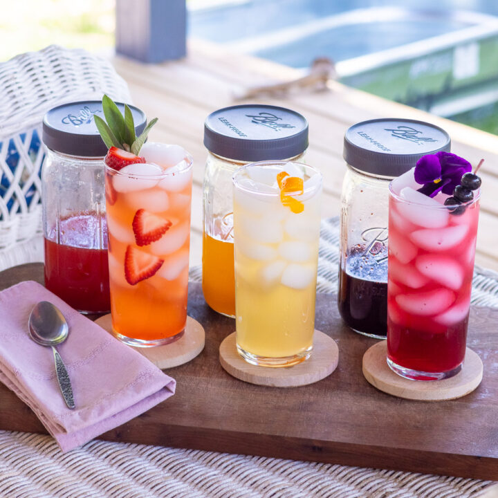Three shrub syrups and garnished drinks on a wooden serving board with a pink napkin and spoon.
