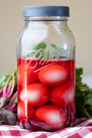 Jar of pickled beets on top of gingham cloth and fresh beets.