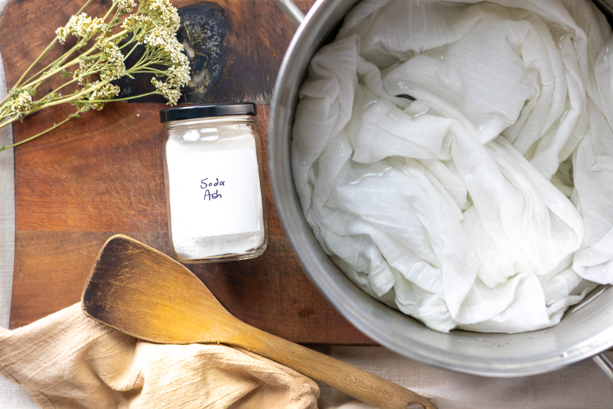 yarrow, soda ash, a pot of white fabric, and a wooden spoon on top of a wooden cutting board.