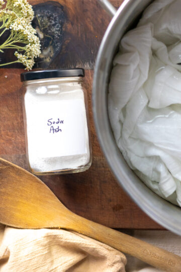 Soda ash, a pot of white fabric, wooden utensils, and dried yarrow displayed in a stylized flat lay.