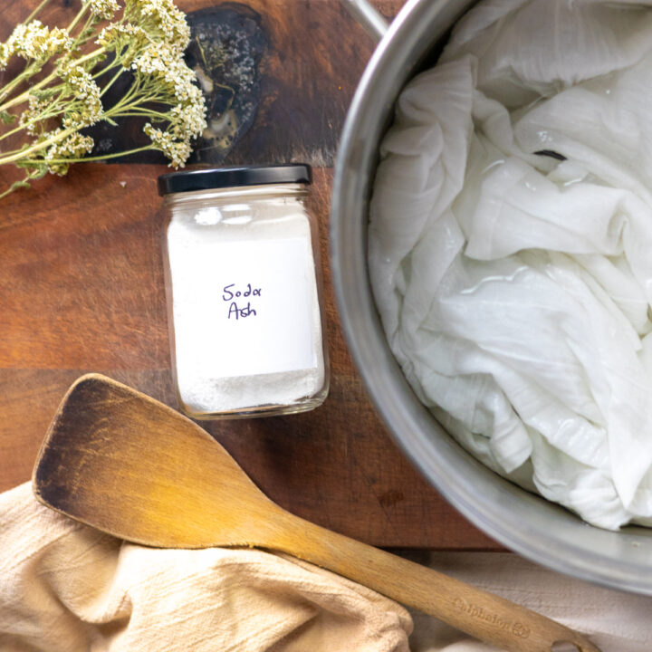 Soda ash, a pot of white fabric, wooden utensils, and dried yarrow displayed in a stylized flat lay.
