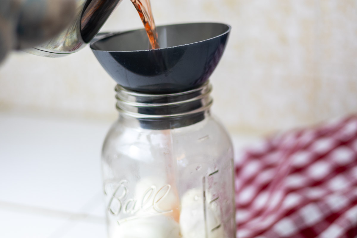 Red brine being poured over hardboiled eggs in jar.