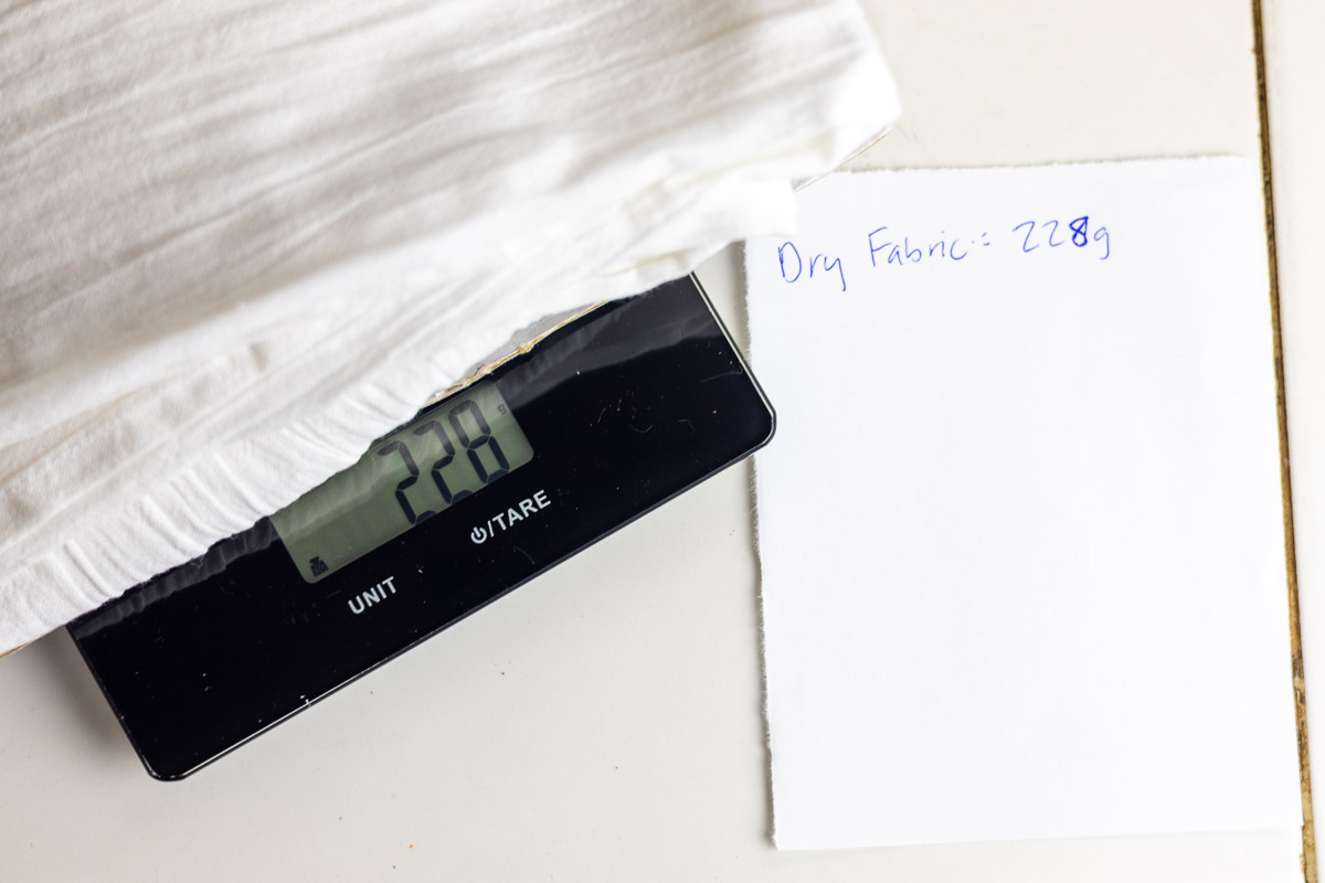 white cotton fabric being weighted on food scale next to scratch paper.