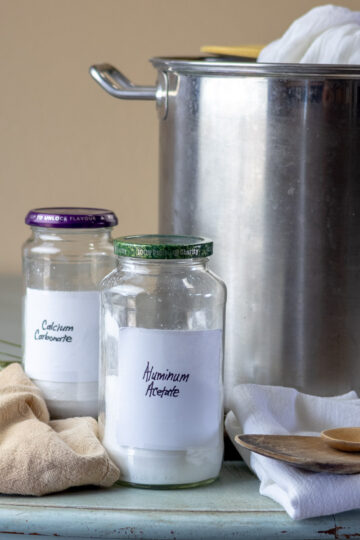 Aluminum acetate mordant ingredients, silver pot, and wooden spoons, layed on a blue table.