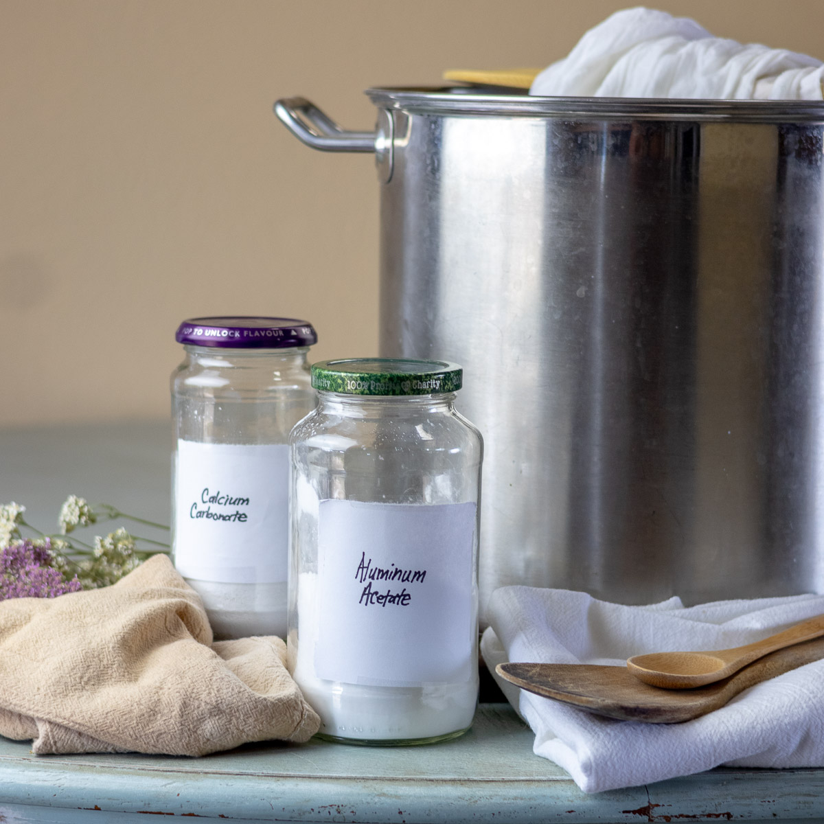 Aluminum acetate mordant ingredients, silver pot, and wooden spoons, layed on a blue table.