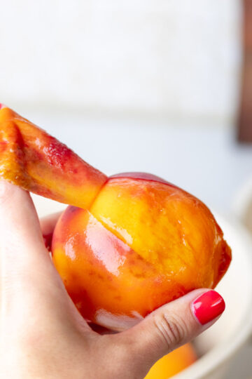 A hand with painted red nails peeling a peach with the index finger.