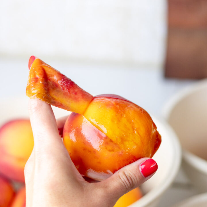 A hand with painted red nails peeling a peach with the index finger.