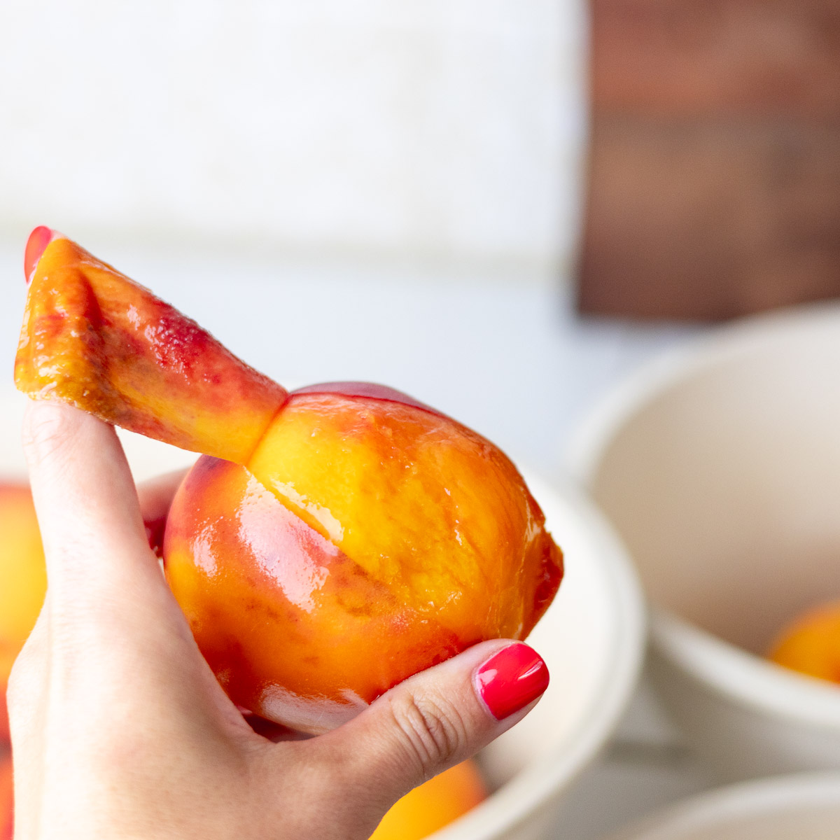 A hand with painted red nails peeling a peach with the index finger.