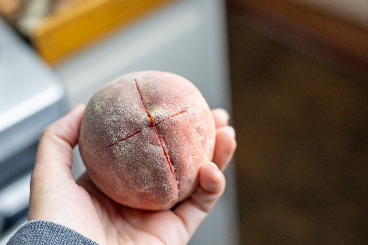 A hand holding a scored peach. An X carved into the bottom of the peach.