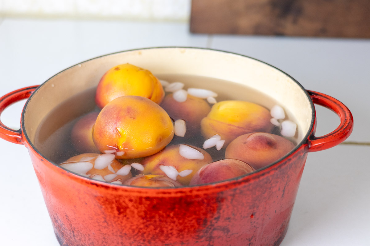 A enameled red cast iron pot filled with ice water and blanched peaches.