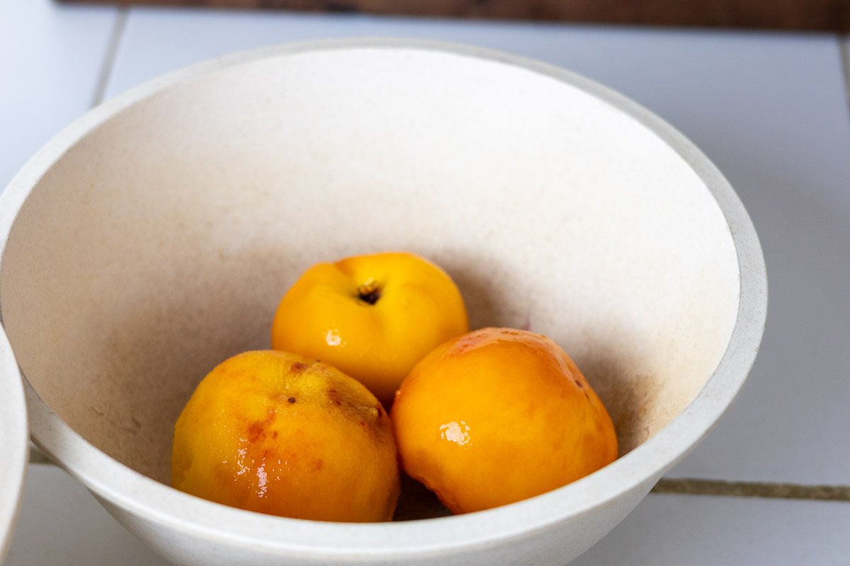 A tan mixing bowl with three peeled peaches.