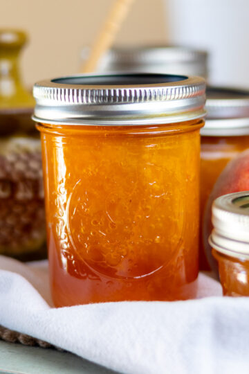 A jar of peach jam styled with white cloth, honey bowl, and peach.