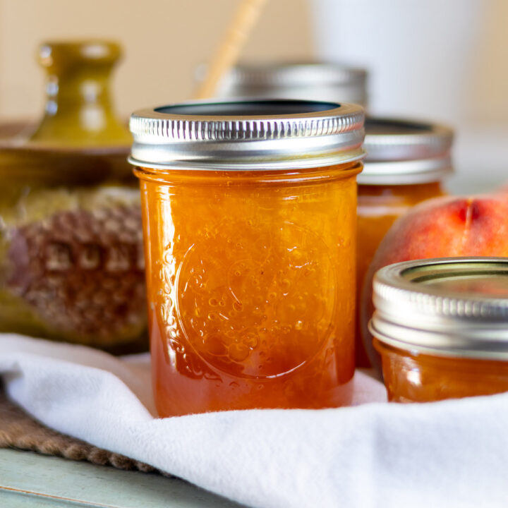 A jar of peach jam styled with white cloth, honey bowl, and peach.