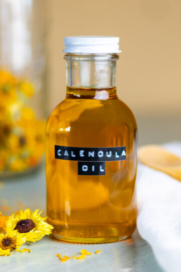 A jar of calendula oil next to dried flowers and petals, and a white cloth and wooden spoon.