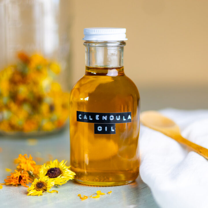 A jar of calendula oil next to dried flowers and petals, and a white cloth and wooden spoon.