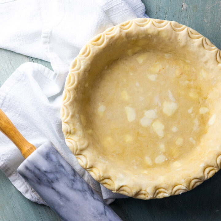 An overhead view of a pie crust with scalloped edges next to a marble rolling pin and white flour sack towel.