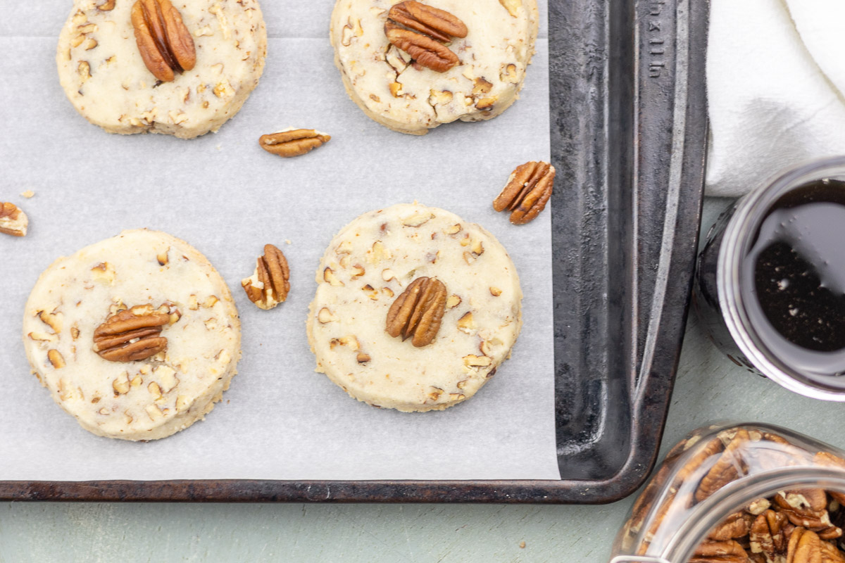 Cookies with pecans on them on a baking tray next to syrup and jar of nuts.