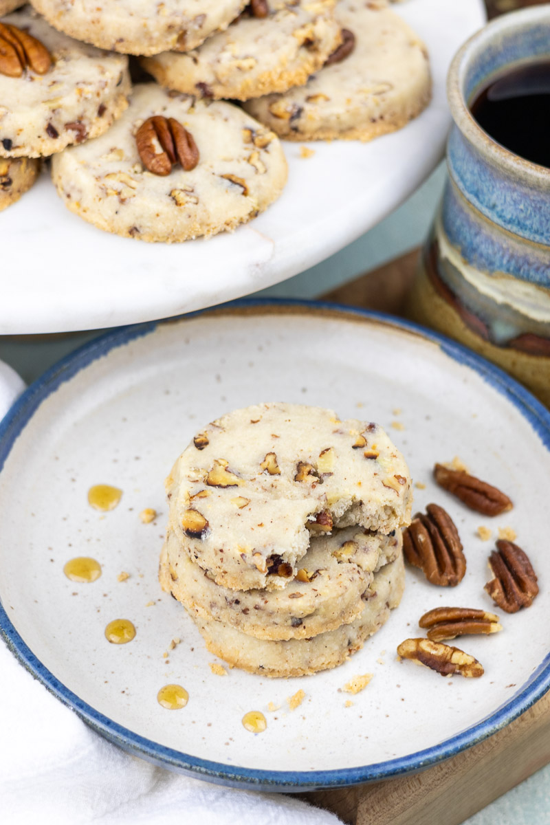 Three maple pecan sandies stacked on a plate next to a cookie tray and coffee.