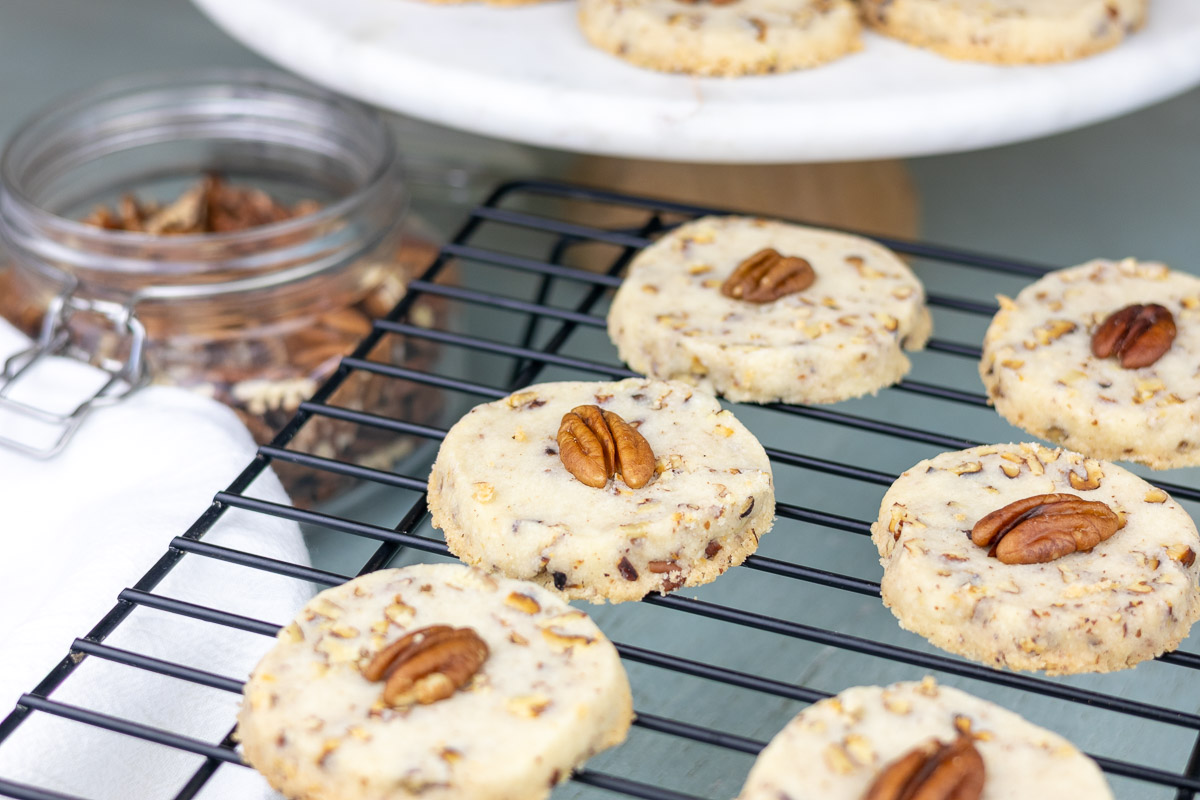A black rack with pecan cookies on them. A jar of nuts and a dessert stand in the background.