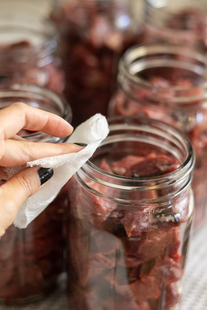 A hand wiping the rims of a mason jar filled with venison chunks for canning raw pack.