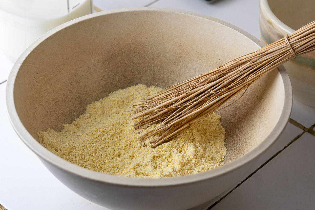 A mixing bowl with cornmeal dry ingredients mixed together with a birch whisk.