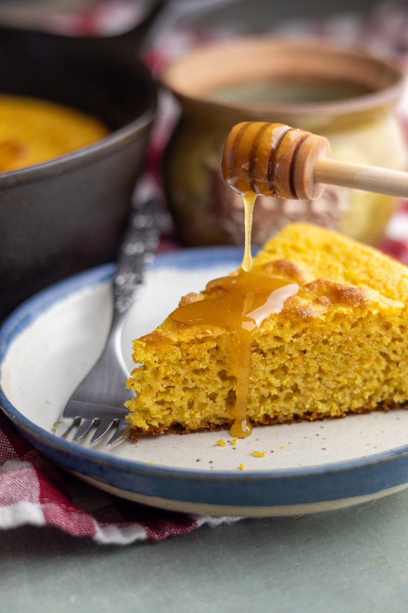 A plate with a slice of cornbread and honey dripping from a honey dipper.