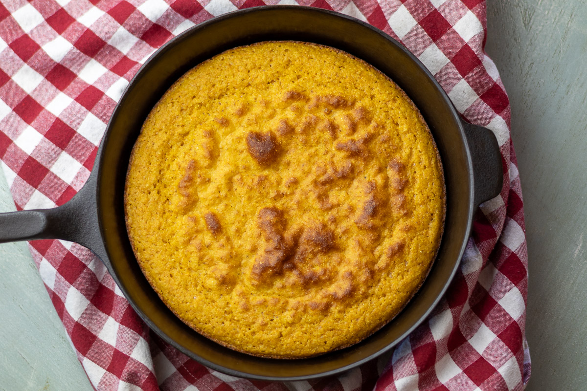 A baked gluten free cornbread in a cast iron skillet on a red gingham cloth.