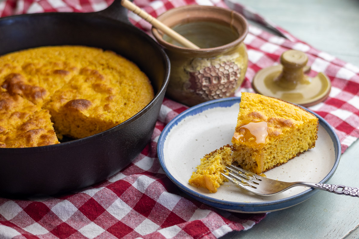 A cast iron skillet next to a slice of cornbread with honey on it.