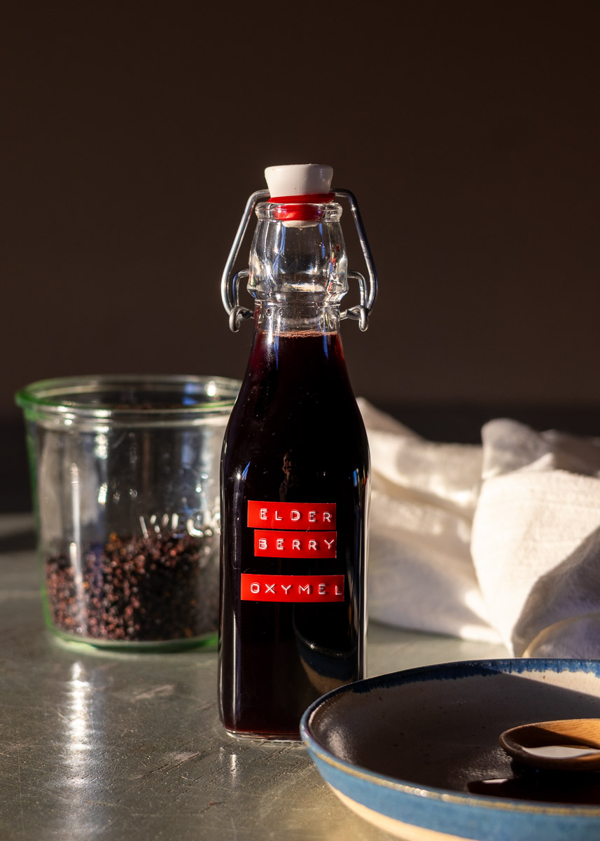 A small swing-top bottle of elder berry oxymel next to a jar of berries and a plate of the syrup.