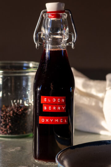 A swing top bottle filled with elderberry oxymel with dry elderberries in foreground and a plate with the oxymel liquid in the front.