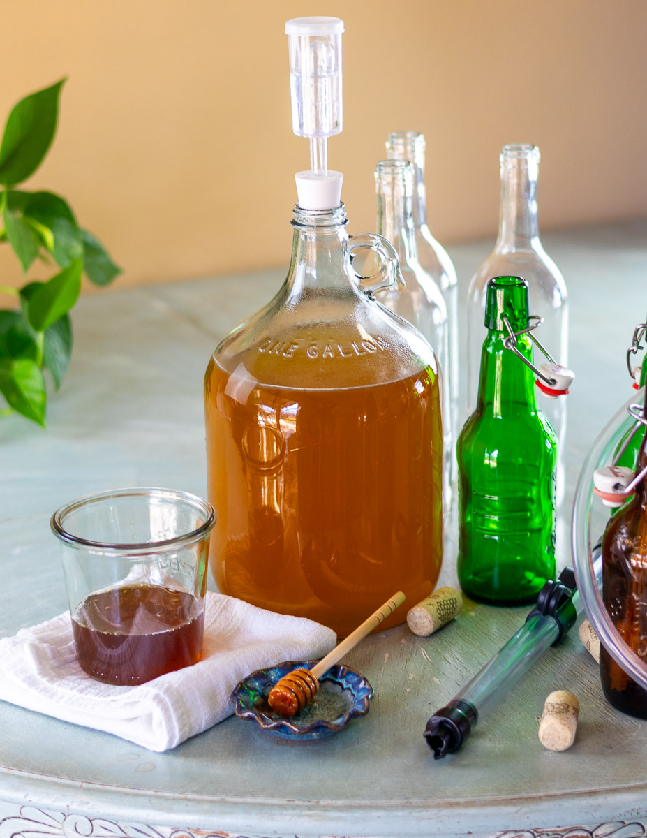 A fermenter with honey mead next to empty bottles, auto siphon, and honey.