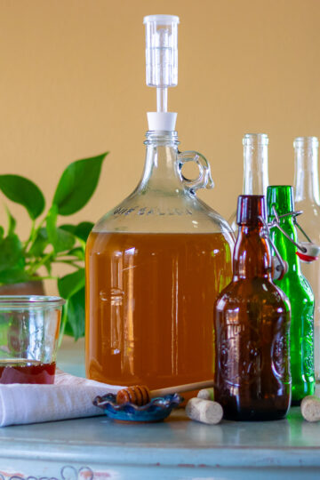 A gallon fermenter of mead next to bottles, honey, and a honey dipper.