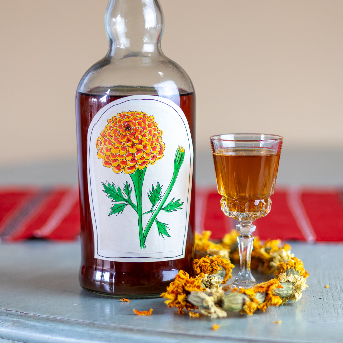 A bottle of marigold liqueur with a hand drawn label, and marigold garland, and cordial glass full liqueur on blue table.